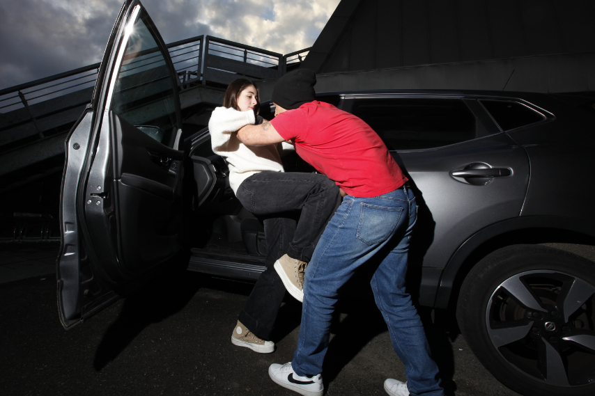 Une jeune femme en pull blanc et pantalon noir se fait agresser à la sortie de sa voiture par un homme portant un tee-shirt rouge, un jean et un bonnet noir.